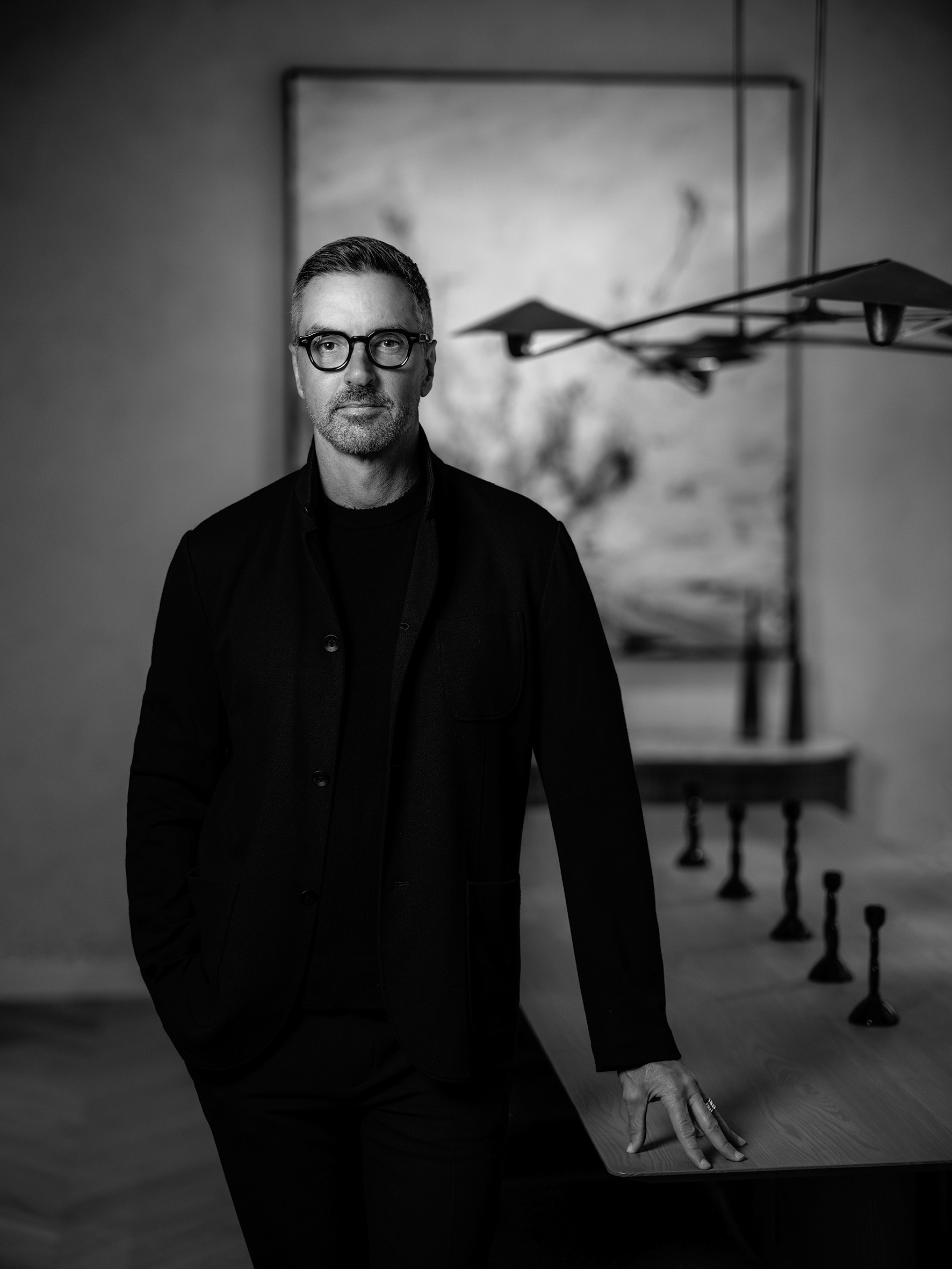 A black and white portrait shot of a middle-aged man with black rounded glasses, a total black outfit, and salt and pepper hair, standing before an abstract painting while leaning on a wooden table.