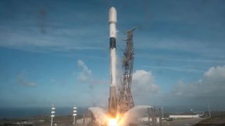A white and black rocket lifts off from its launch pad into a partially cloudy blue sky.