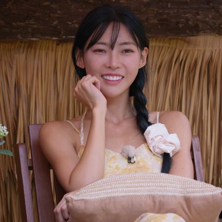 A woman (Kim Min-gee) wearing a yellow dress and a white scrunchie around her hair braid. She sits with a pillow on her lap, in front of a straw wall and next to white flowers in a vase, in the Netflix Korean reality show 'Single's Inferno' season 5.