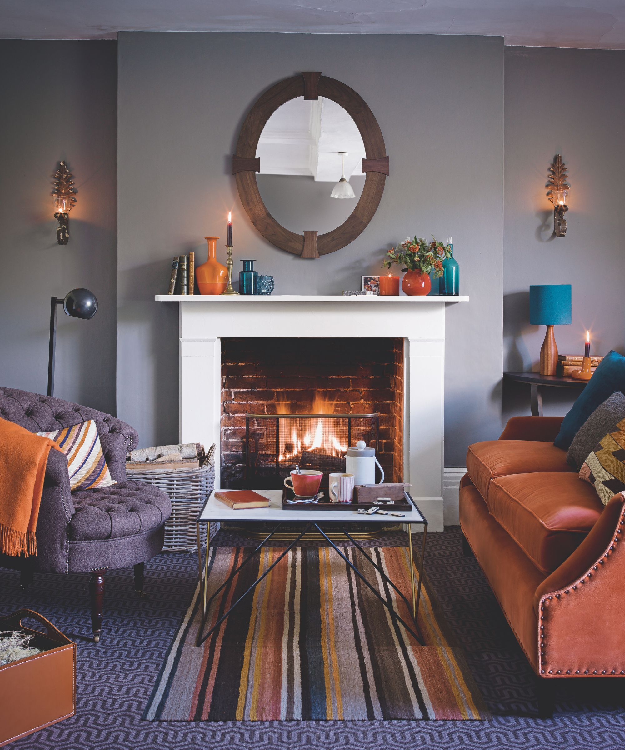 A lilac living room with a white mantelpiece, orange and purple chairs and sofas with matching cushions, and a striped rug underneath a coffee table in the center of the room.
