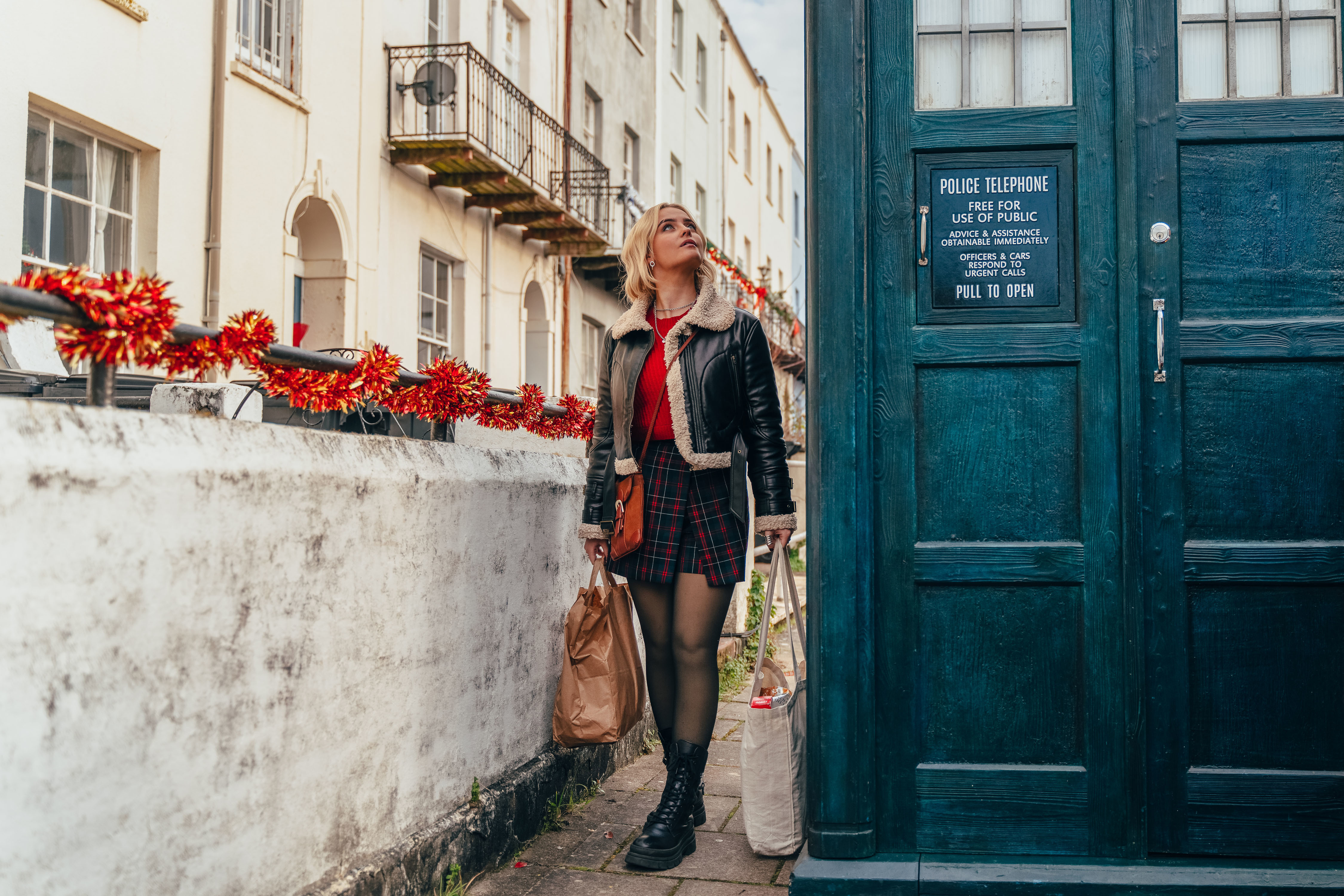 Ruby Sunday (Millie Gibson) is walking down a residential street carrying a bag of shopping in each hand. The Tardis is right next to her on the pavement and she is looking up at it with curiosity.