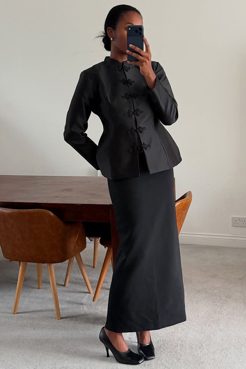 A woman taking a selfie in a mirror wearing a black tang jacket styled with a long black pencil skirt, black high-vamp square toe pumps, and diamond earrings.