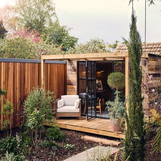 A wooden garden room with cream upholstered chair and Crittal-style black framed doors