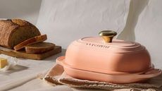 A pink cast iron oval bread oven on a folded dish towel, with a loaf of fresh sliced bread in the background. 