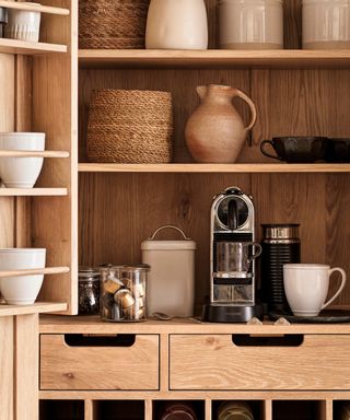 The inside of a wooden pantry with shelving. A coffee maker on the bottom shelf, pots and baskets on the shelves above.