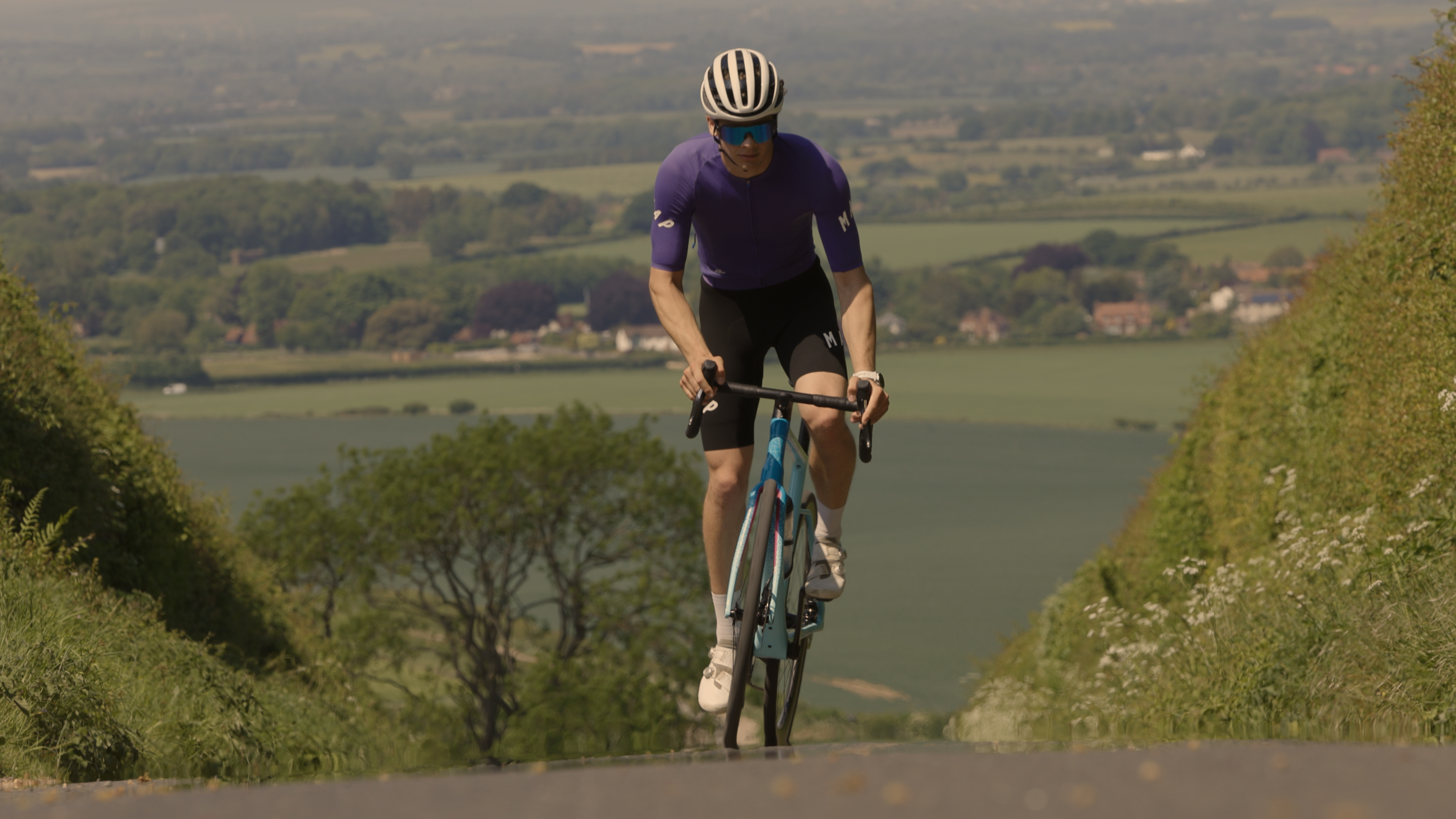 A cyclist is riding a road bike in a purple jersey and black shorts on a lane with countryside in the background