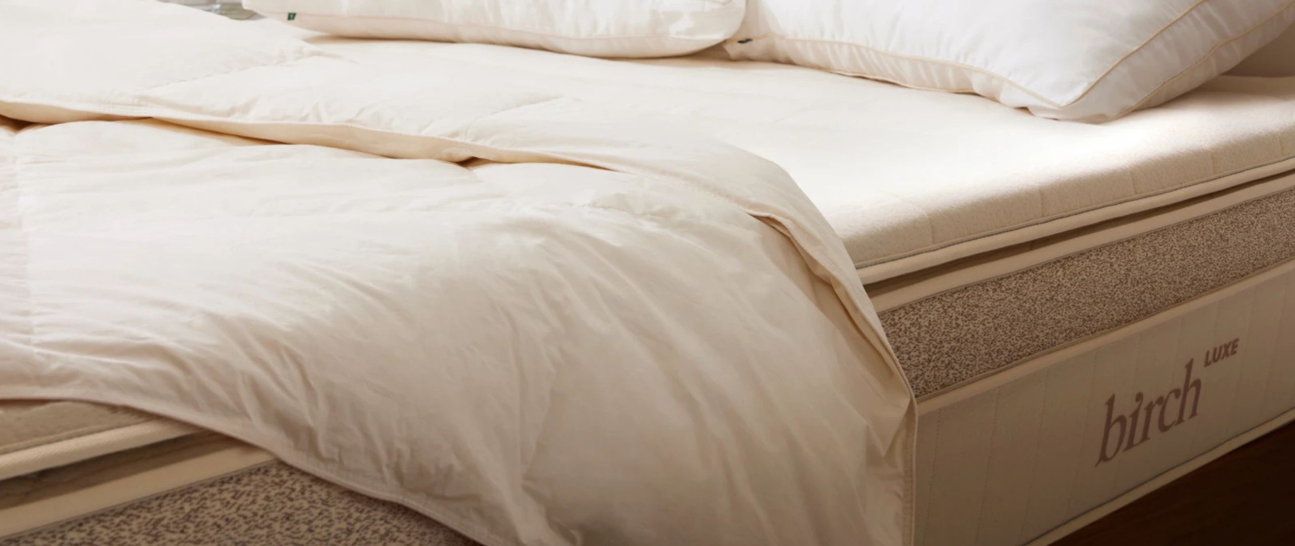 A close up of the Birch Plush Organic Mattress Topper placed on top of a Birch latex mattress with a off-white comforter draped over it. 