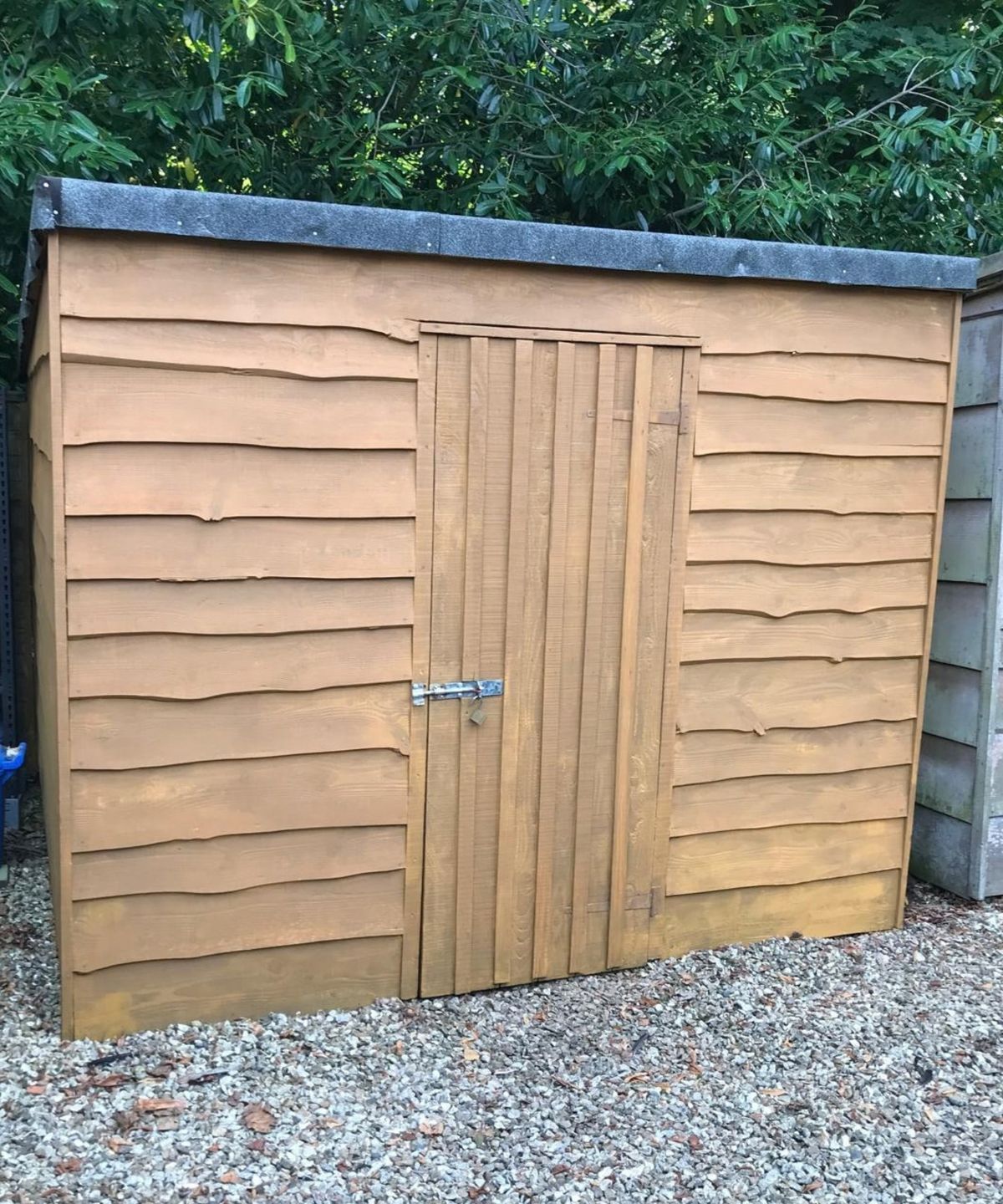 Wooden garden shed with a felt roof, latched door, and overlapping cladding, set on gravel.