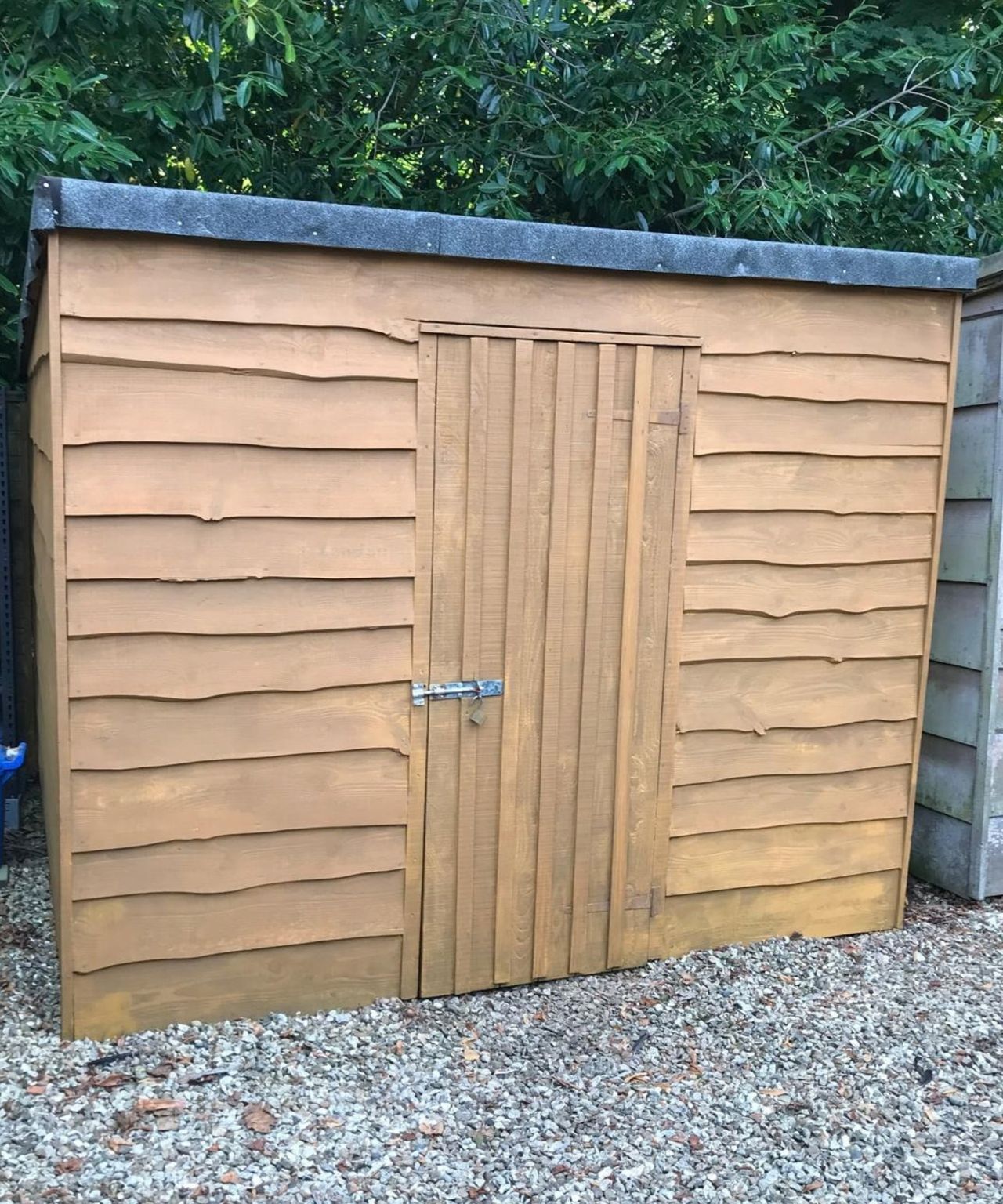Wooden garden shed with a felt roof, latched door, and overlapping cladding, set on gravel.