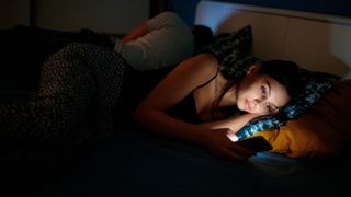 A woman with dark hair lays in bed in a dark bedroom while scrolling on her phone 