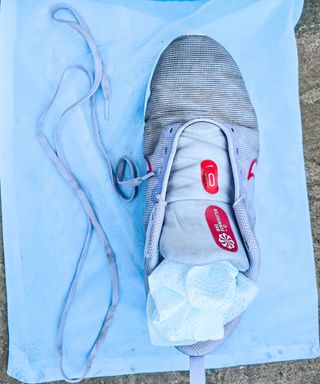 A purple and red trainer with white tissue inside of it and purple laces next to it, on top of a white mesh bag