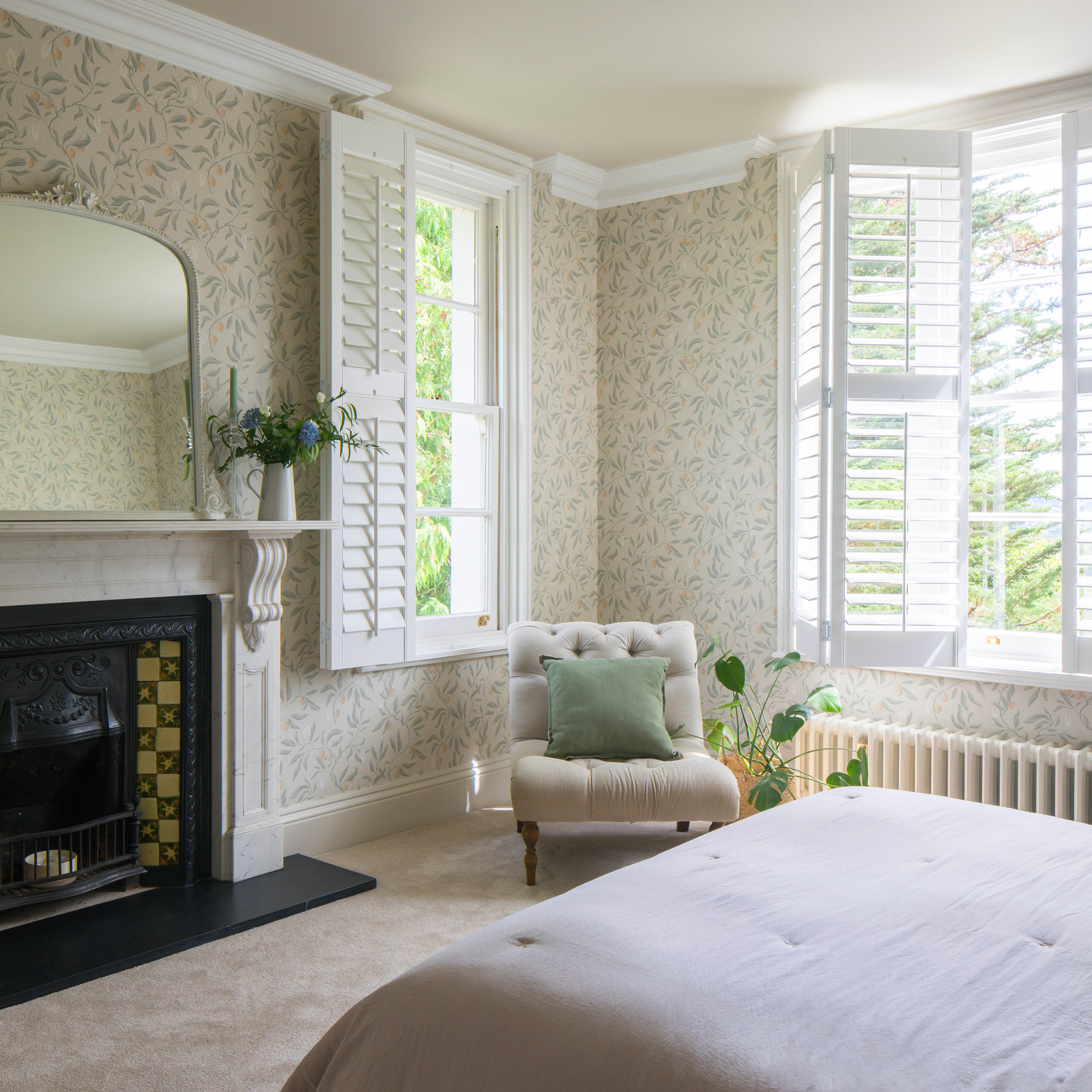 a bedroom with botanical wallpaper on the walls, a traditional fireplace and two windows with shutters