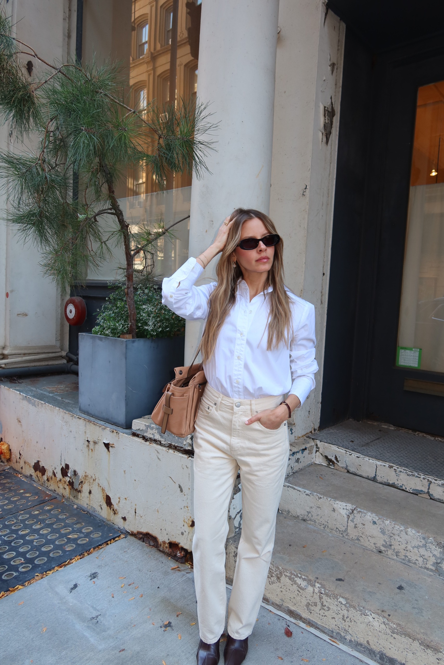 Nikki wears a white button-down shirt, ivory jeans, brown boots, and a tan bag.