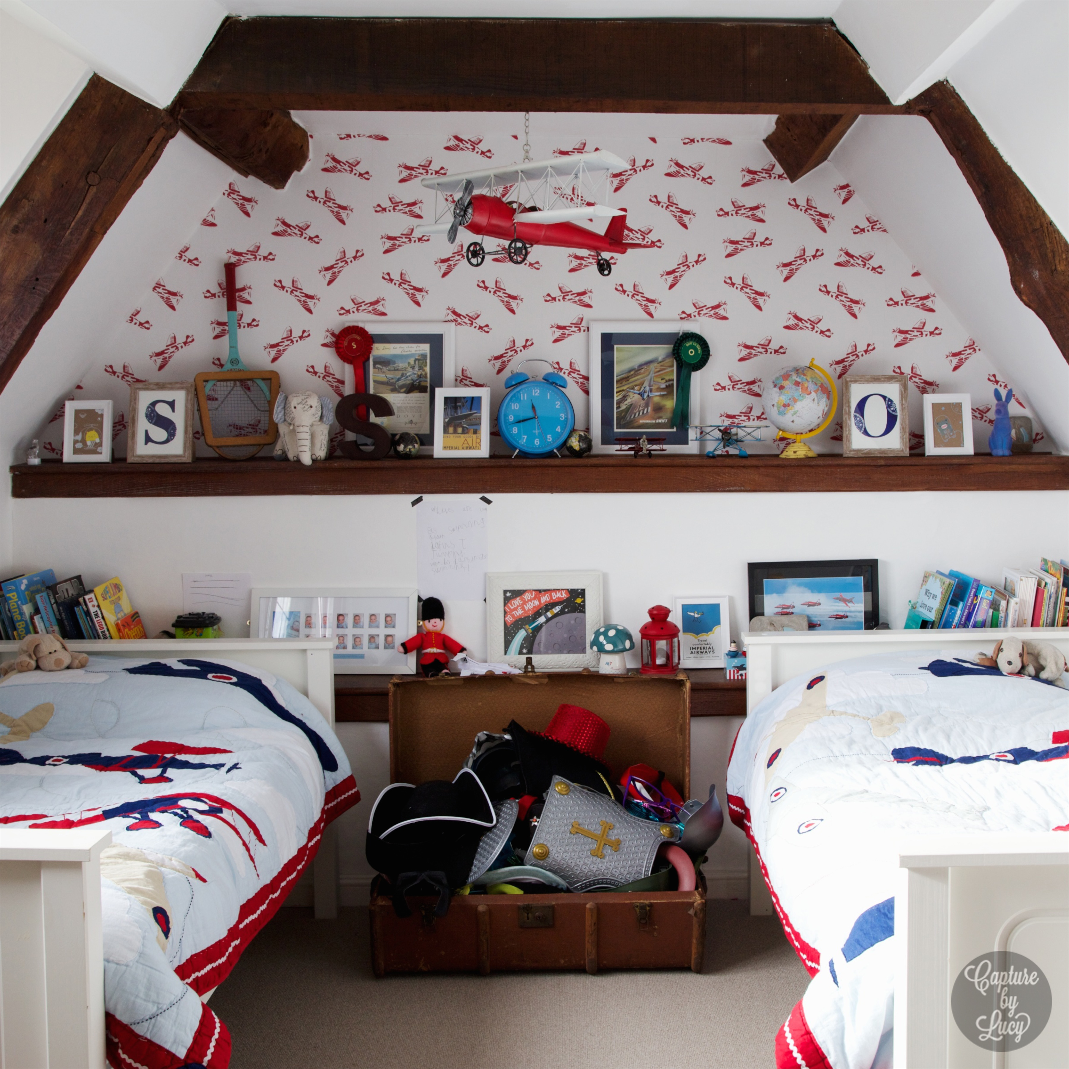 Attic shared bedroom ideas featuring a red aeroplane wallpaper, blankets and toy hanging from ceiling, built-in wooden shelving and a storage trunk.