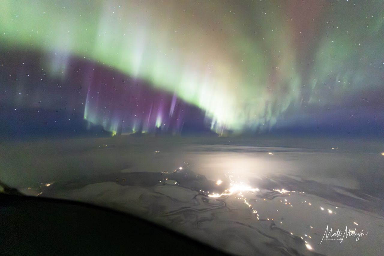 curtains of green auroras fill the sky, with broken clouds below and distant glows of city lights.