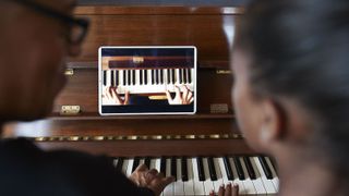 Two people sat at a piano watching a lesson on a tablet