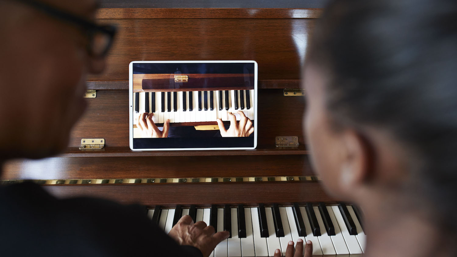 two people sitting at the piano watching the text on the tablet