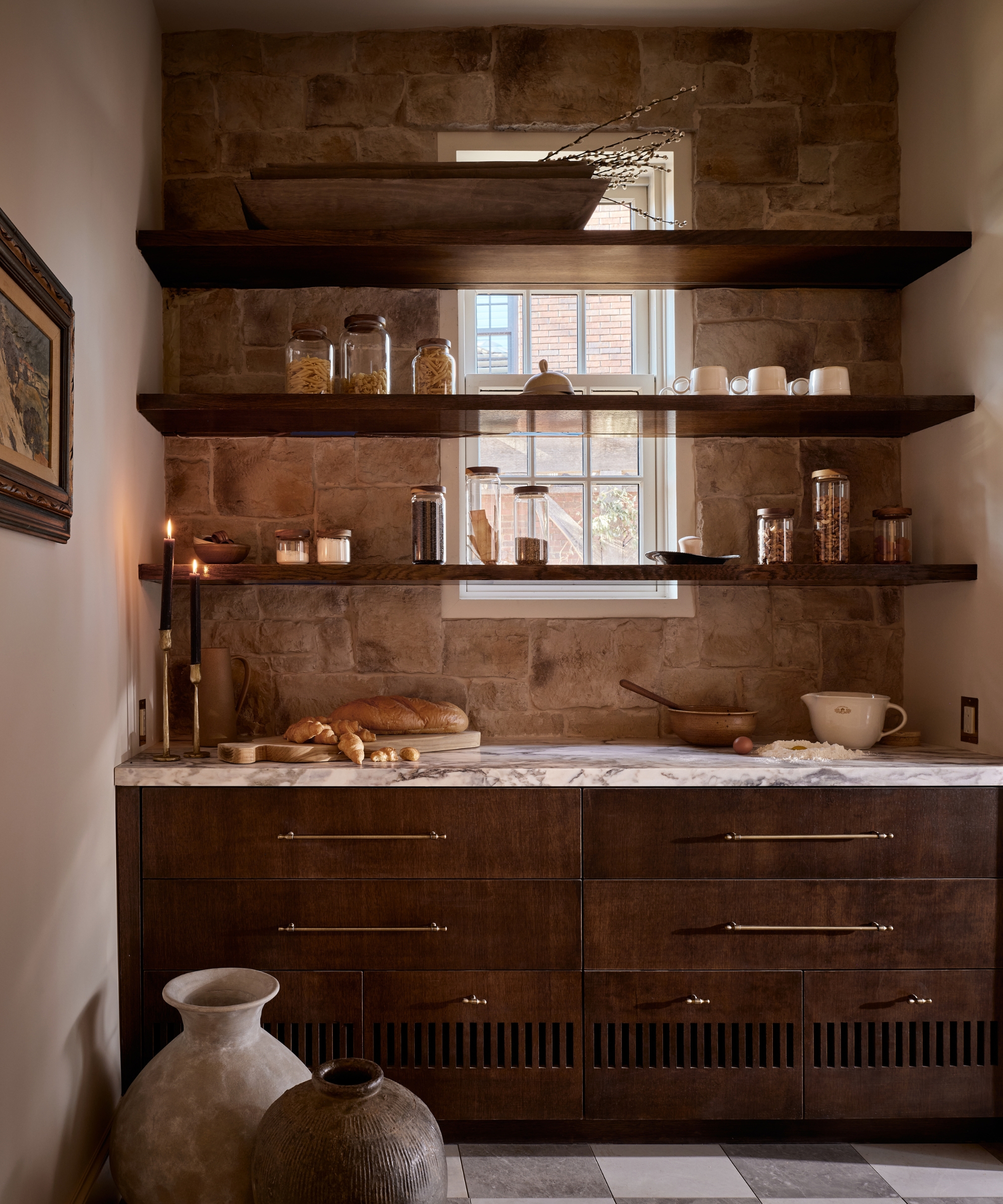 A walk-in pantry with exposed stone walls, open shelving that goes over the window, wooden cabinets, and marble countertops