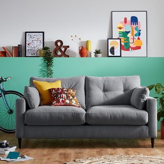 A grey sofa with yellow and orange cushions in a green living room with a wall shelf of pictures and ornaments