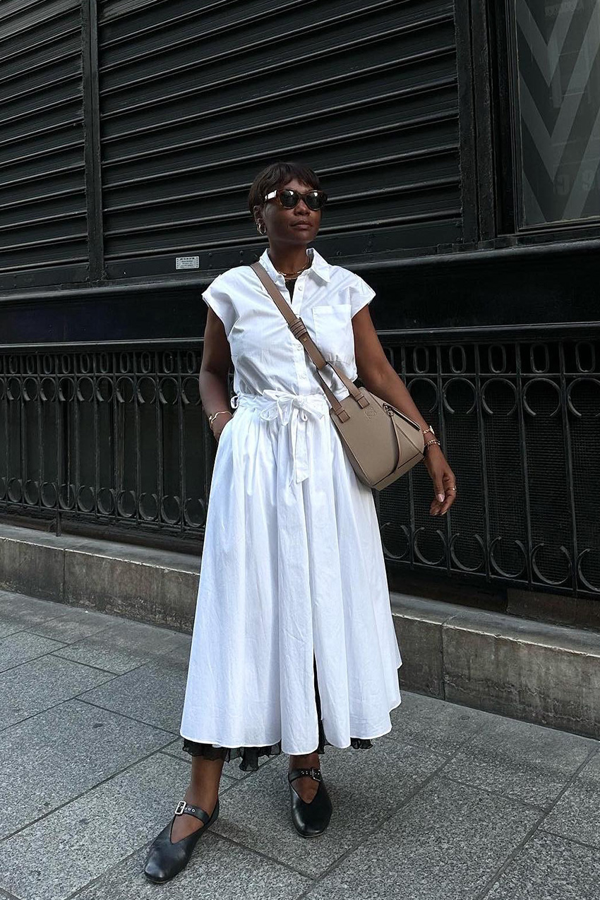 summer airport outfit is shown in a street style photo of a French woman wearing brown sunglasses, gold hoop earrings, a white shirt dress, ballet falts, and a Loewe crossbody bag