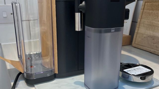 Side view of the Nespresso Vertuo Lattissima, showing the water tank, milk carafe and silver cup rest/drip tray. This pic is before assembly so there are still some packing elements present.