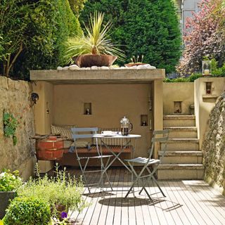 Patio area with metal garden table and chairs seating area in alcove concrete stairs leading to house ledge with large plater trees