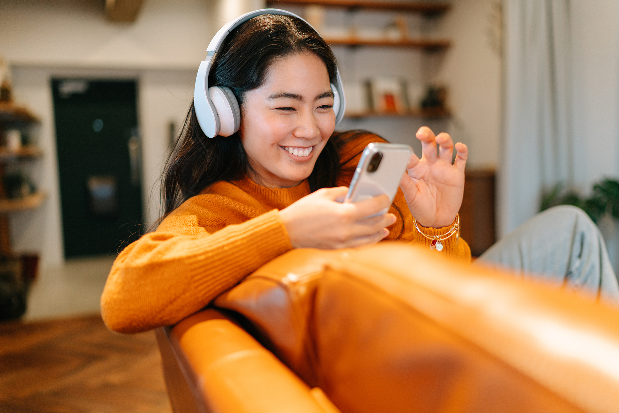 A woman in headphones sits on a leather sofa looking at a phone
