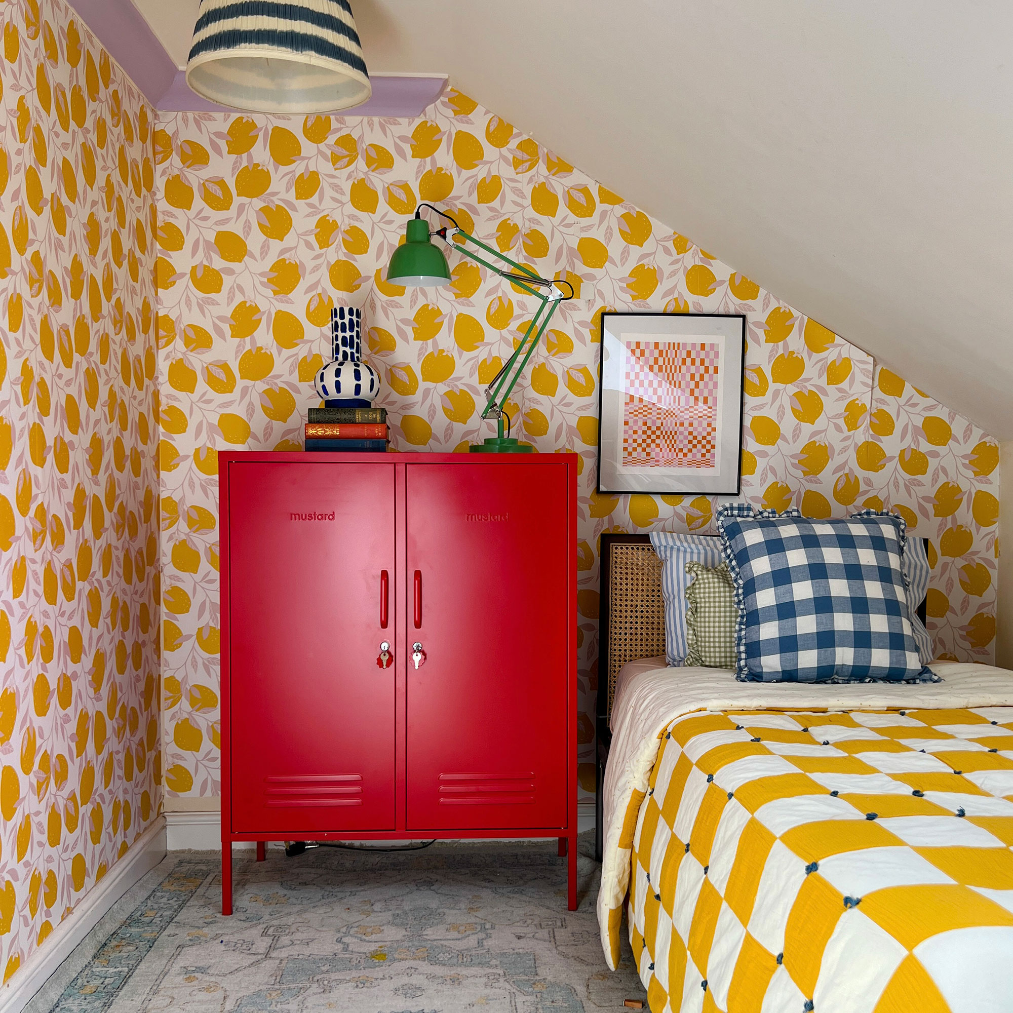 Eaves bedroom with yellow patterned wallpaper, and chequered yellow bedding. Red locker cabinet used as bedside table with green desk light