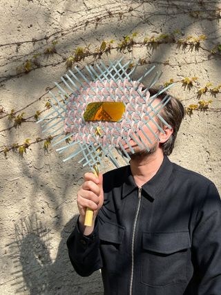 A portrait of German designer Sebastian Herkner whose face is hidden by a round fan woven with pink and light blue cord