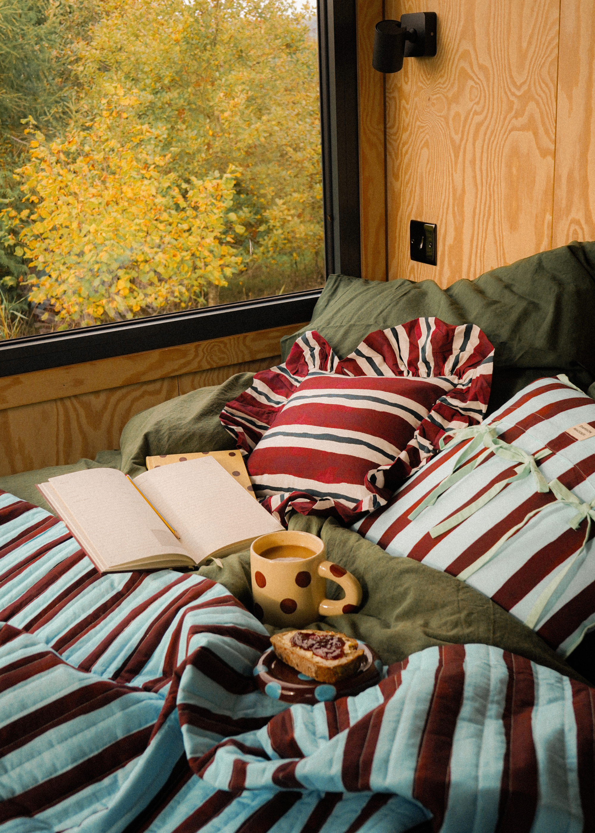 A wood cabin with a green bed featuring striped pillows, a ceramic mug, a diary, and a plate with a slice of toast and jam