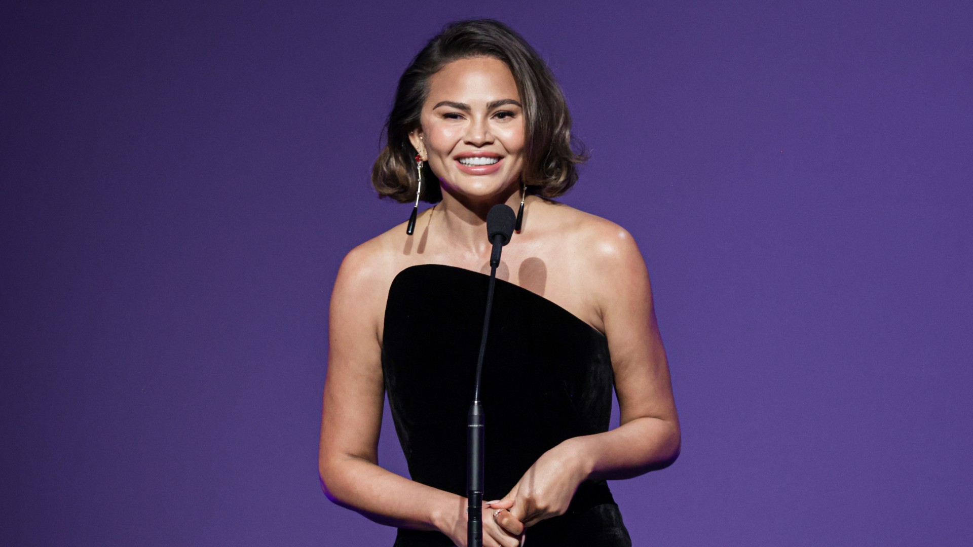 chrissy teigen speaks on stage wearing a black and white gown