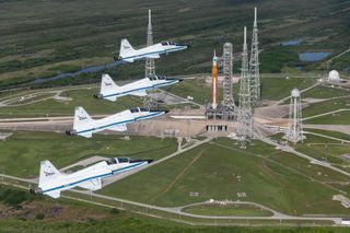 four white jets fly above a large orange rocket standing on a launch pad