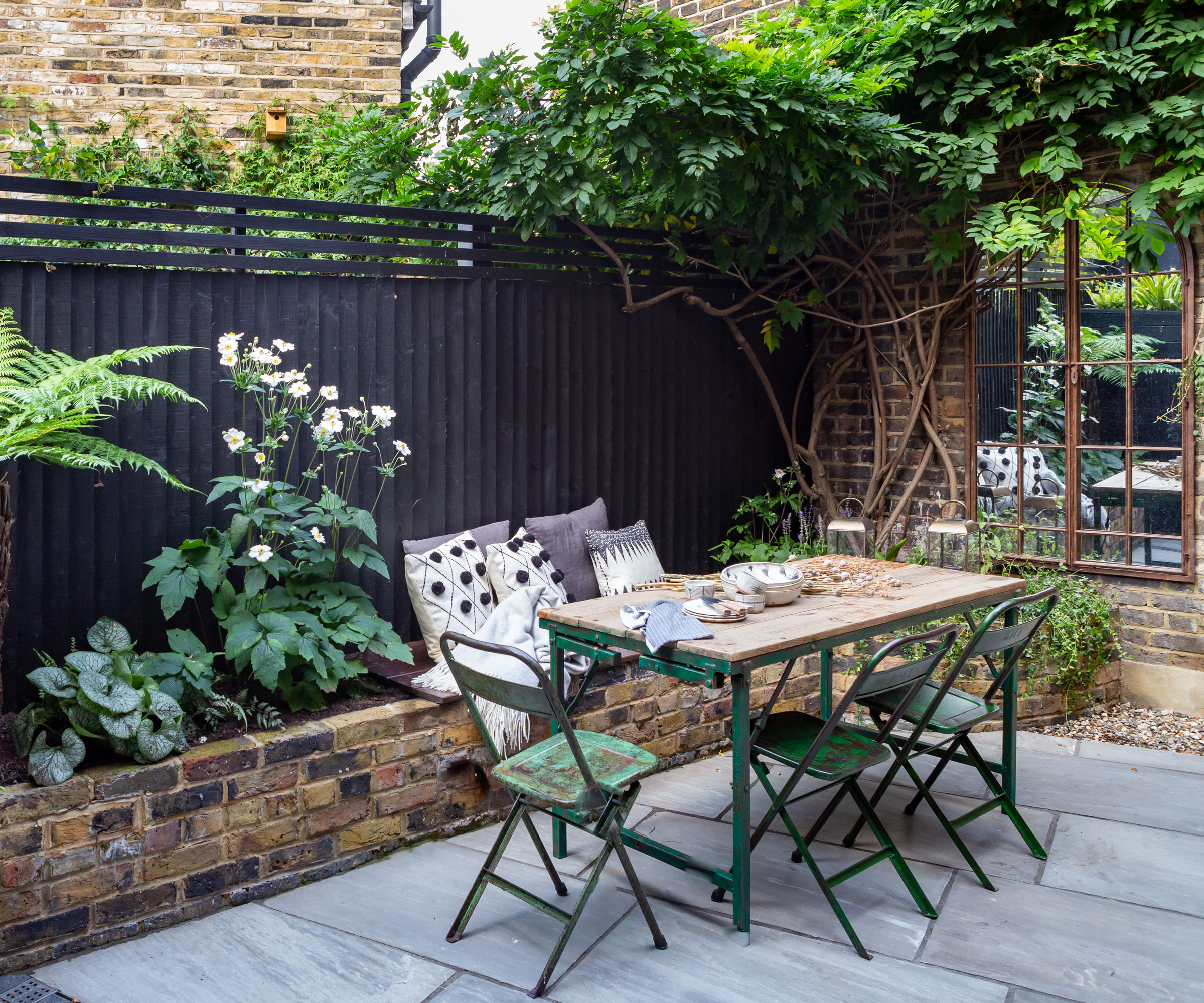 small patio with dining table set, mirror, black fence, raised beds with planting and climbing plants