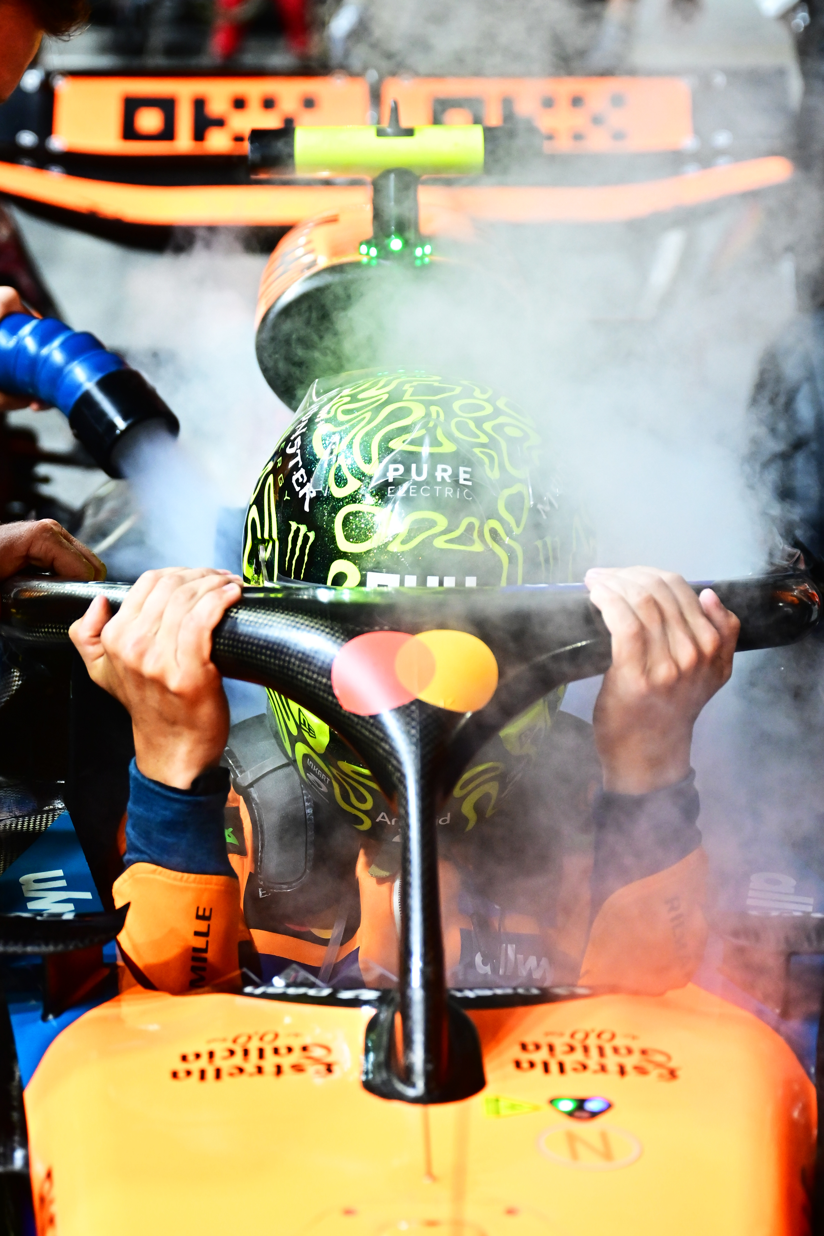 Lando Norris lowers himself into his F1 car while holding onto the Halo. He&#039;s in an orange race suit and a black and neon yellow helmet