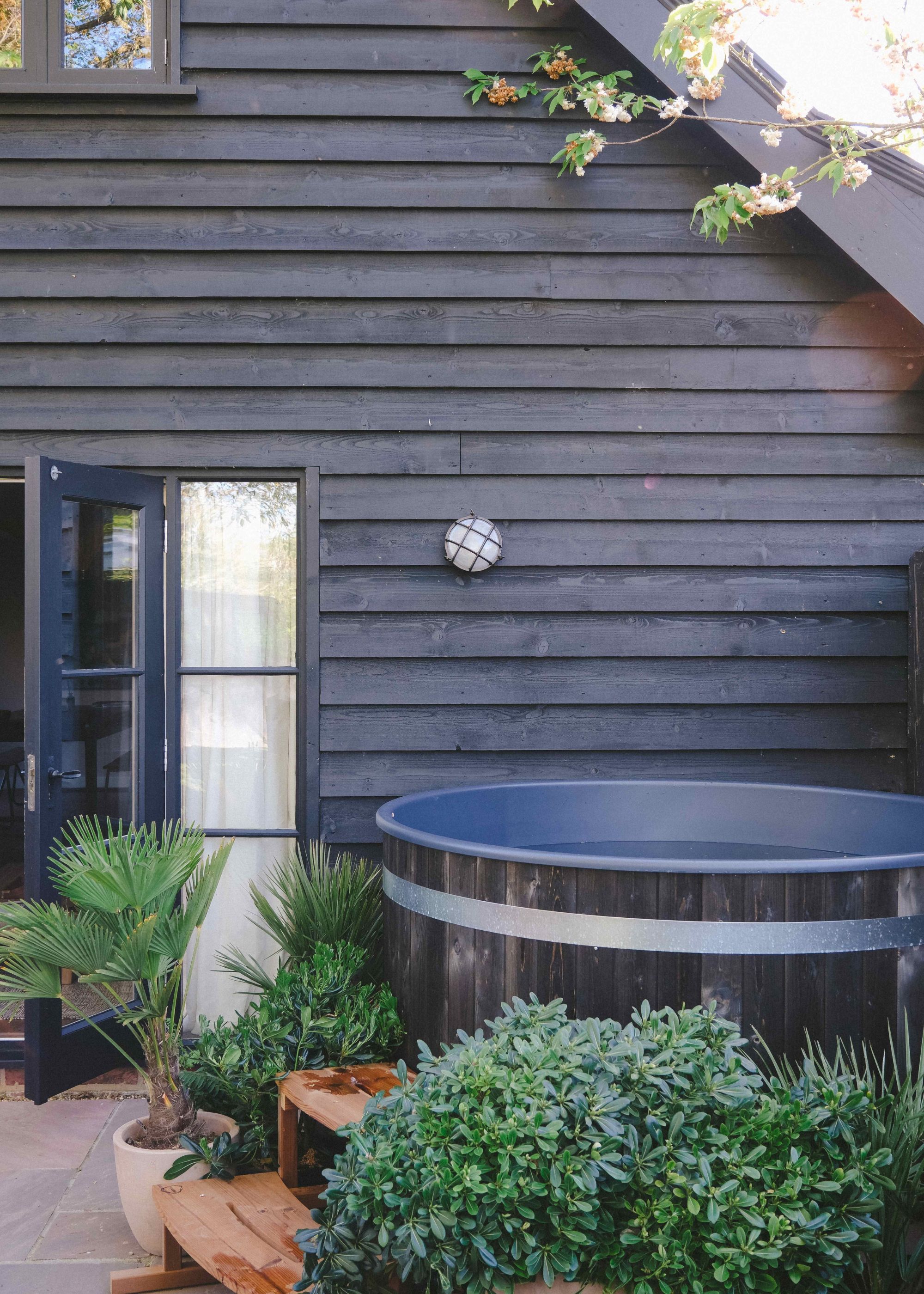 A home exterior with dark gray wood-clad walls, a sloped roof, glass doors with a black frame, and a patio with a hot tub.