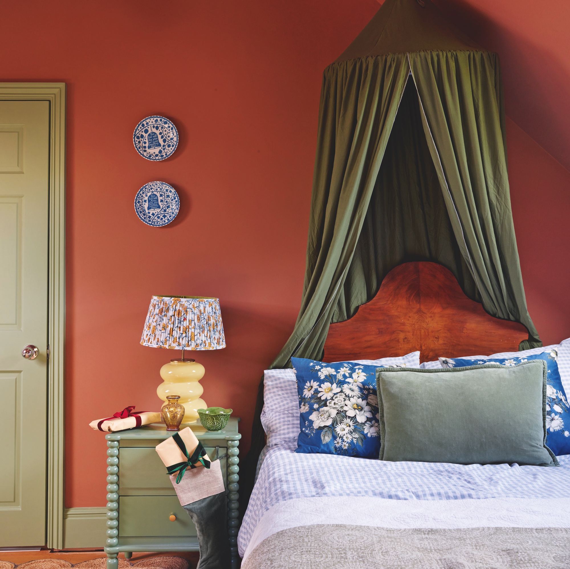 bedroom with orange walls and green canopy above single bed with wooden headboard and blue floral cushions, and green bobbin bedside table