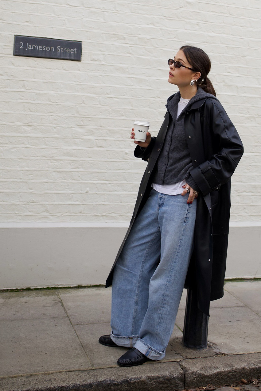 Denim and shoe combos shown in a photo of a street style photo of a woman standing outside on the sidewalk wearing a long black leather jacket on top of a gray sweatshirt and white t-shirt styled with wide-leg cuffed jeans, black loafers, silver hoop earrings, and black sunglasses