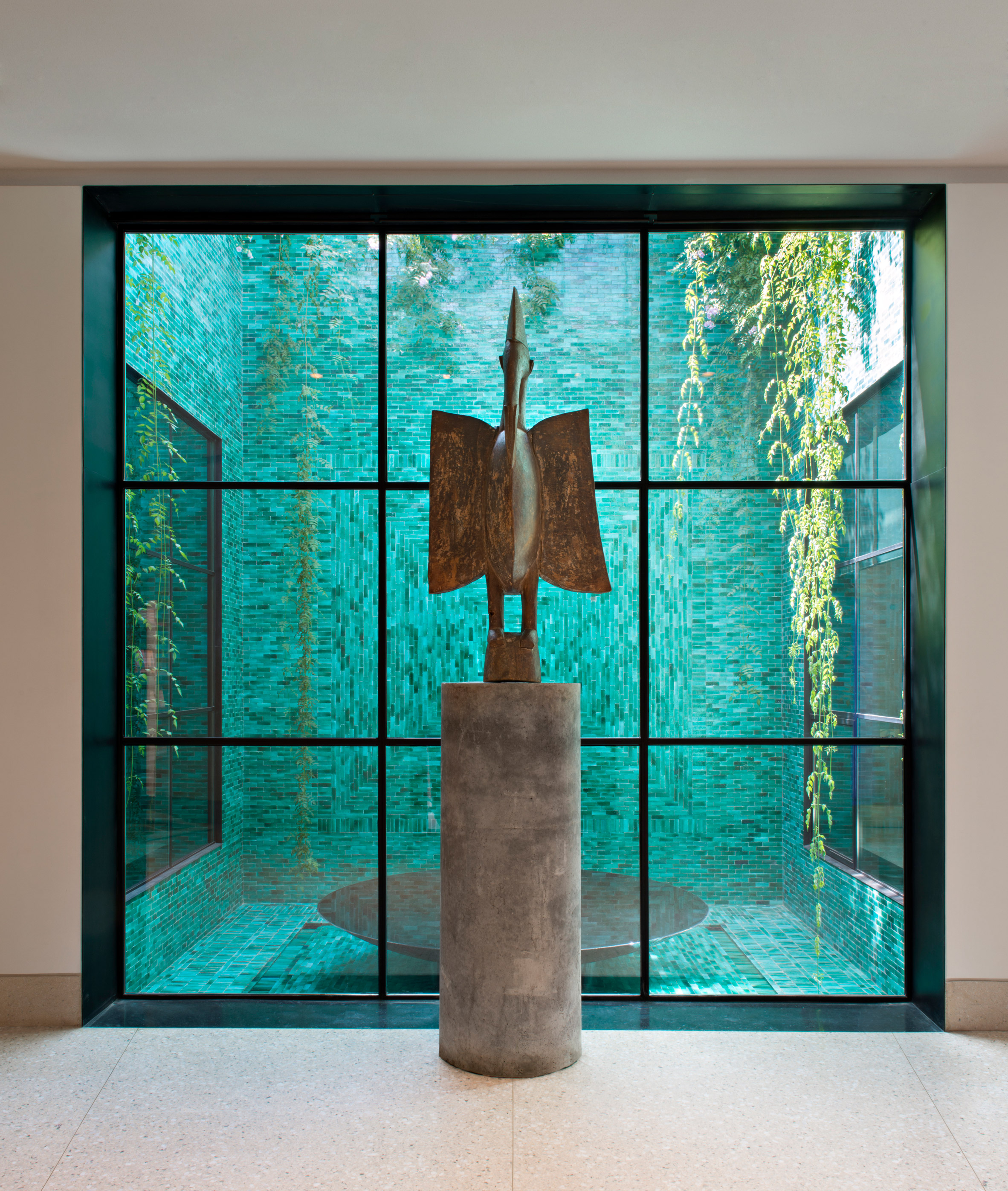 A museum gallery room features a bird-shaped sculpture in bronze set against a glass window outside of which lies a seagreen-tiled courtyard with plants and a round, silver fountain.