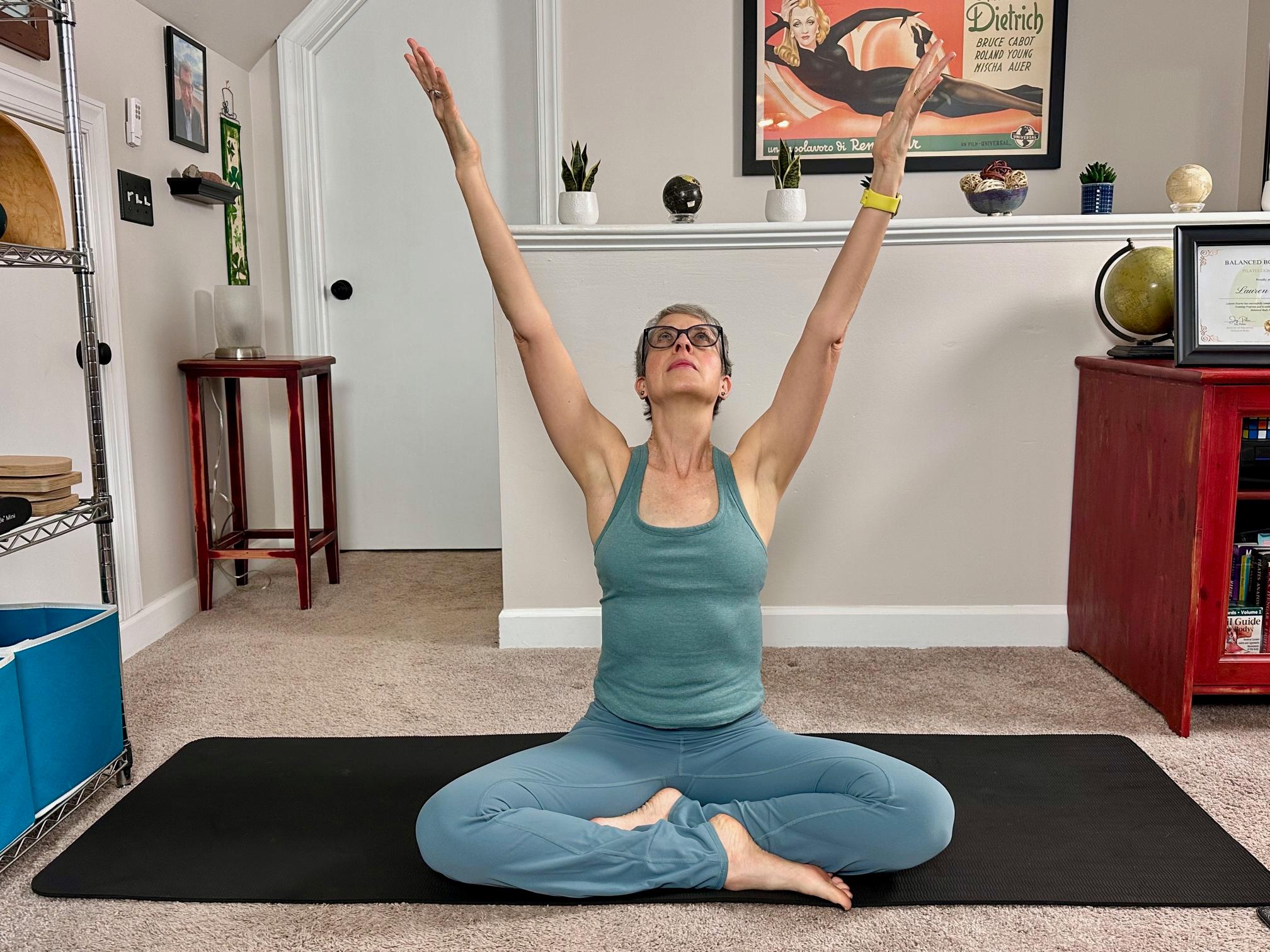 woman sitting cross-legged reaching her arms upward on a black yoga mat in a living room