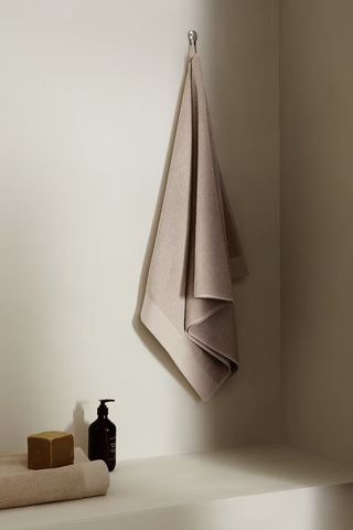 neutral bath towel hanging on wall with folded towel, hand pump, and cake of soap next to it