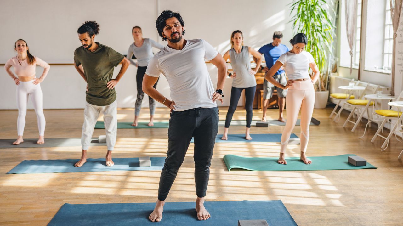 A group of people perform hip circles on yoga mats in a brightly lit exercise studio. They have their hands on their hips and they are bending to the side. We see a large plant at the back of the studio. 