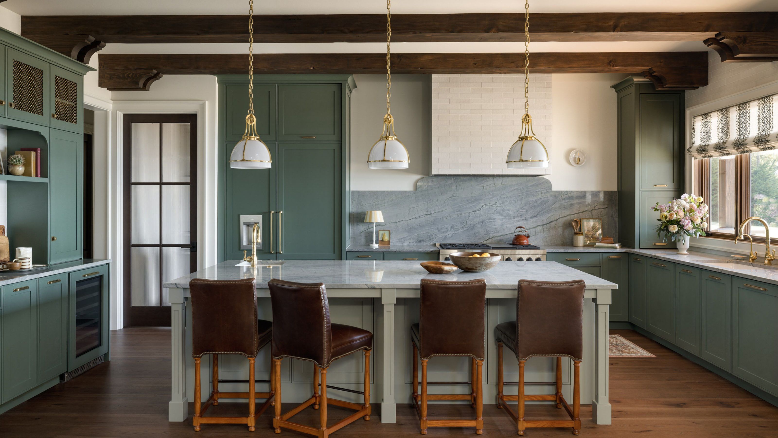 A large kitchen with muted green cabinets, a white island, wood flooring, warm white walls, and a ceiling with dark wood beams.