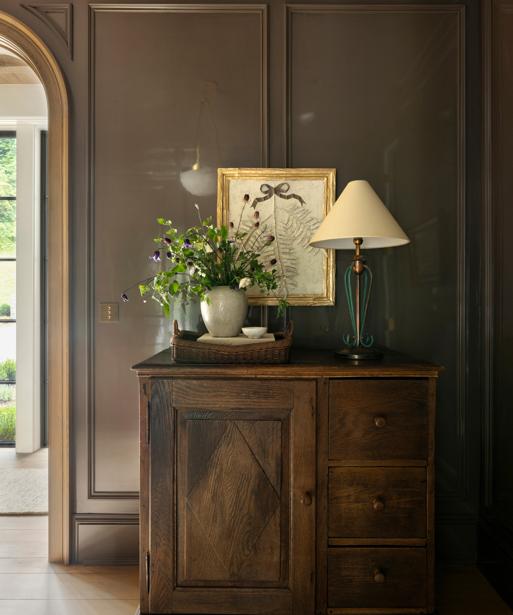 Entryway with dark brown walls, vintage wooden console with rattan tray and vase of flowers on top and a vintage metal lamp