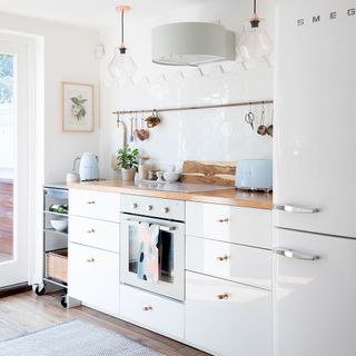 All white kitchen with wooden work surfaces, white wall tiles and copper rail hanging with utensils