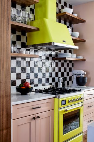 kitchen with timber cabinetry, black and white checkerboard backsplash, neon yellow stove and range hood, stand mixer on the counter, and bowl of fruit, with floating timber shelves on either side styled with glasses and plates