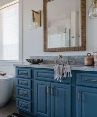 A bathroom with a brown woven large mirror, a gold wall sconce, a blue sink unit with cabinets and drawers and a gray marble surface, gold taps, two oval wooden storage holders and a gray seashell trinket holder