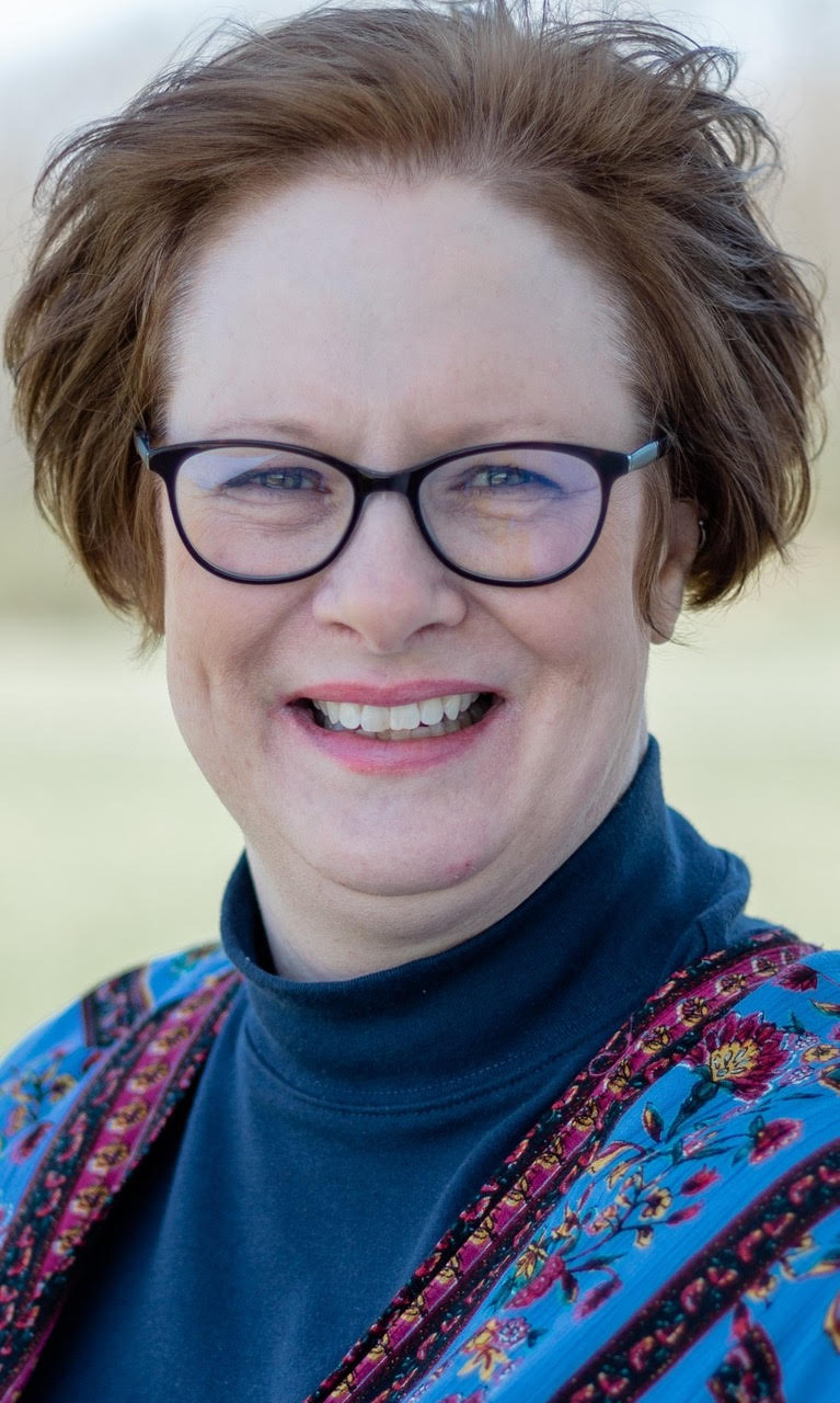 Headshot of woman with short brown hair, glasses, wearing a navy turtleneck top