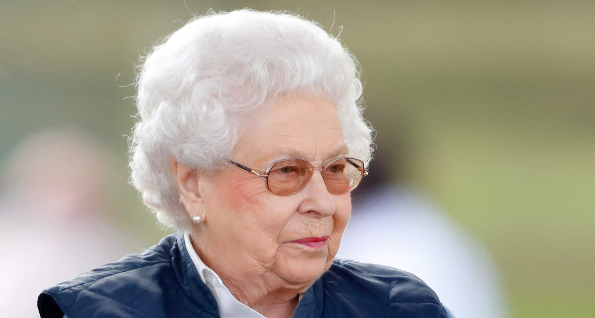 Queen Elizabeth wearing a blue vest and glasses and smirking