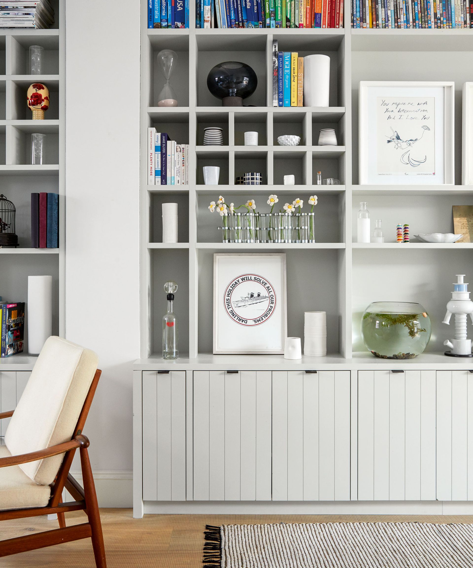 nbuilt storage cupboards and shelves in pale grey with books and ornaments beside wooden flooring and a grey rug.