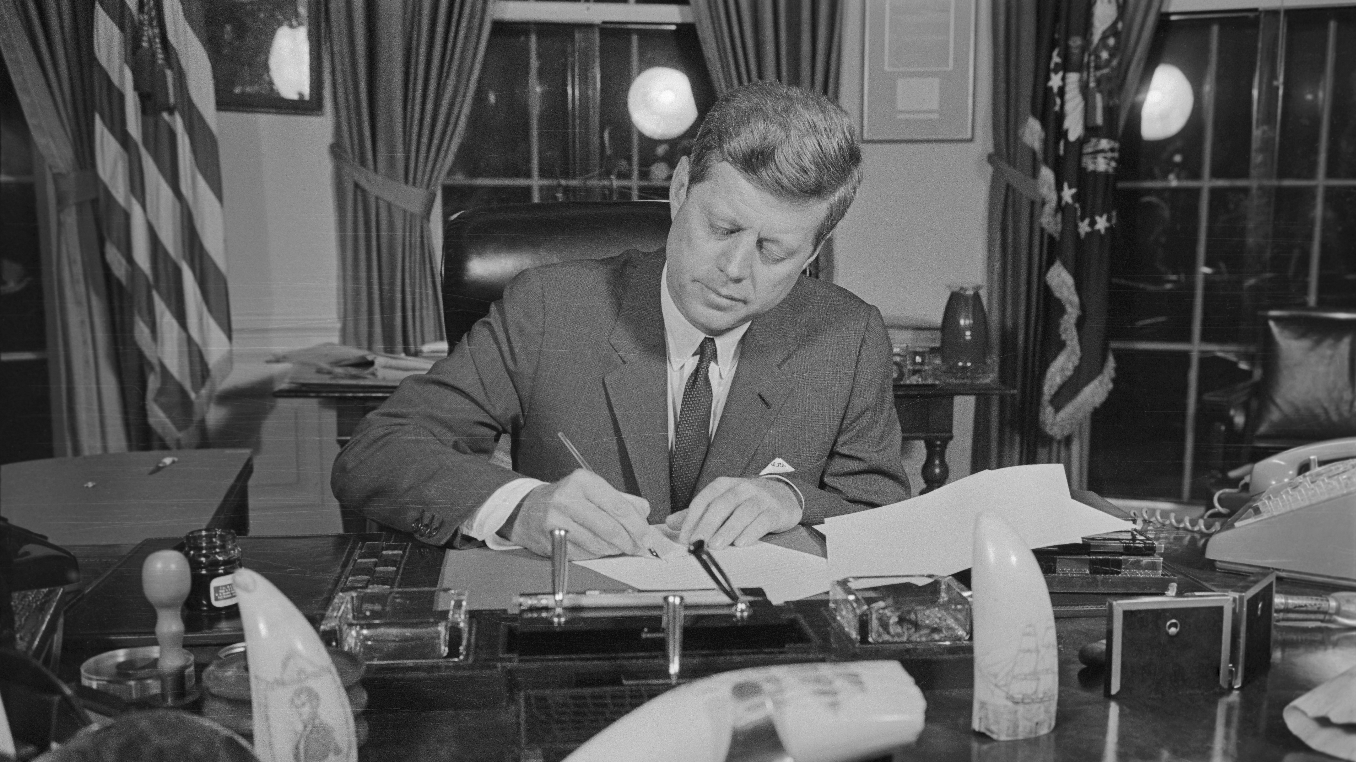 President John F. Kennedy sitting at his desk in the oval office signing a US arms quarantine against Cuba.